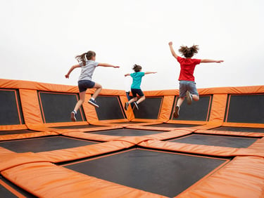 Low-angle wide shot of kids jumping on wall-to-wall trampolines, bright high-key lighting, energetic orange padding, crisp action-freezing details