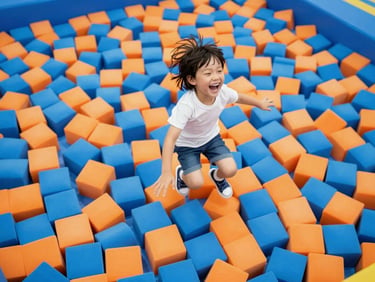 A child laughing mid-air as they leap into a giant pit filled with soft blue and orange foam cubes, bright high-key lighting