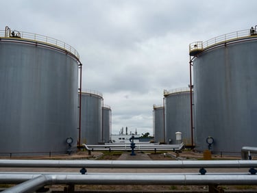 Wide shot of a petrochemical storage tank farm, large cylindrical tanks receding into the distance under cool overcast sky, access pipework in the foreground, institutional muted grey and navy tones