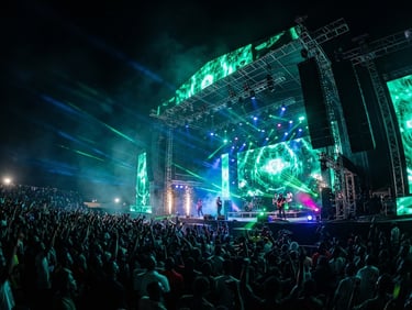 Low-angle wide shot of a massive concert stage at night in Benin, towering giant LED screens displaying vibrant green graphics, professional line-array sound systems, dramatic emerald and blue lasers cutting through light haze, grand scale.
