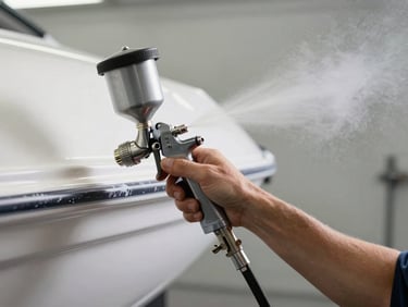 Technician's hand holding a spray gun applying marine topcoat to a boat hull, fine mist visible in north-facing workshop light, clean neutral background, no faces