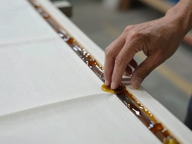 Close-up of a hand pressing fiberglass cloth into a hull joint, resin glistening under shop tungsten light, structural laminate layers visible at the edge
