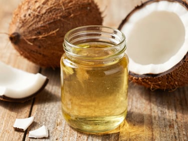 warm editorial photography, natural side-lit sunlight, rich golden and amber tones, shallow depth of field, 35mm, close-up of clear wood-pressed coconut oil in a clean glass jar, whole coconuts and dried coconut flakes on a rustic wooden surface in the background.