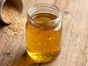 warm editorial photography, natural side-lit sunlight, rich golden and amber tones, shallow depth of field, 35mm, close-up of amber wood-pressed sesame oil in a clean glass jar, raw sesame seeds on a rustic wooden surface in the background.