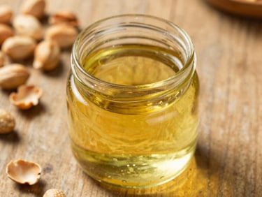 warm editorial photography, natural side-lit sunlight, rich golden and amber tones, shallow depth of field, 35mm, close-up of golden wood-pressed groundnut oil in a clean glass jar, raw groundnuts and their shells on a rustic wooden surface in the background.
