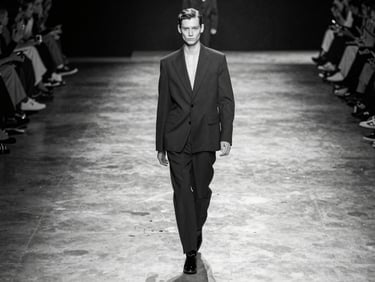 Stark black-and-white wide shot of a model walking a minimalist concrete runway, harsh direct overhead lighting, sharp dramatic shadows, raw high-fashion atmosphere.