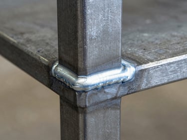 Extreme close-up, studio strobe lighting — a steel table leg-to-apron weld joint, cool white light raking across the weld seam to reveal the bead texture and clean grind finish, warm concrete floor visible softly out of focus in the lower background, no props, no accessories