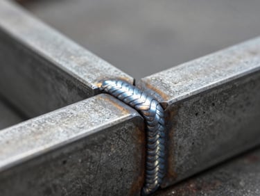 Close-up of welded steel joint where two flat steel bars meet at right angle, showing the finished weld bead in sharp detail, soft north-facing studio light revealing the surface texture and the precision of the joinery, shallow depth of field, neutral gray background, emphasis on the craft of the connection