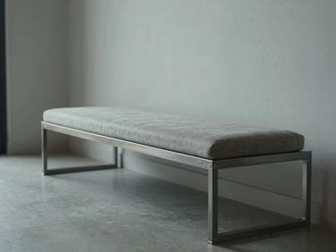 Wide panoramic of a steel bench with upholstered seat in a minimal residential entryway, cool north daylight, steel frame with visible joinery, one hand resting on the frame, tactile detail visible, generous negative space