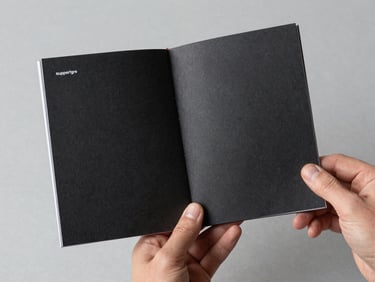 A hand holding a freshly printed, saddle-stitched zine with heavy textured black cover and minimalist typography