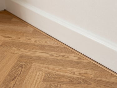 Close-up detail of freshly laid oak parquet flooring meeting a newly painted baseboard, natural side light revealing wood grain texture, no people, tight framing