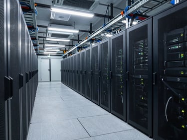 Wide shot of a data center floor with rows of server racks and visible overhead cable trays and cooling units, cool fluorescent lighting, no people