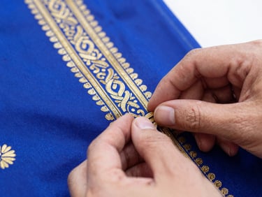 Macro close-up of hands carefully inspecting intricate gold embroidery on a royal blue silk saree, bright natural daylight, crisp detail, 50mm lens