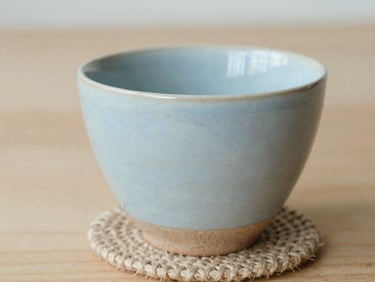 Macro close-up of a hand-thrown ceramic cup with raw clay texture and a soft pastel blue glaze, sitting on a jute coaster, minimalist composition, warm daylight