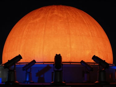 A wide environmental shot of a planetarium dome lit in deep solar orange, silhouetted telescopes in the foreground.