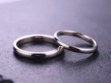 Macro shot of three minimalist 925 silver rings catching sharp light on a dark reflective charcoal surface, soft lavender background glow