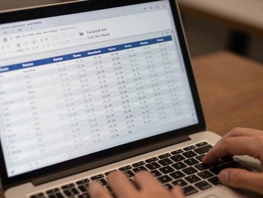 Close-up of a spreadsheet on a laptop screen showing Facebook Ads cost-per-result columns, a hand resting near the trackpad, neutral overhead office light, numbers clearly legible