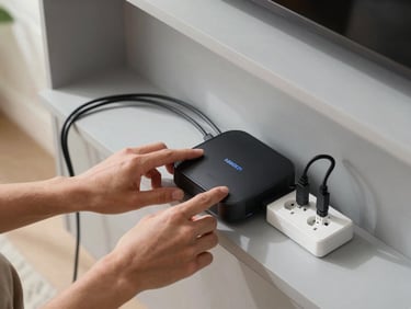 Overhead view of hands connecting a smart home hub to a power socket, device cables neatly arranged on a light grey shelf, natural daylight from the left