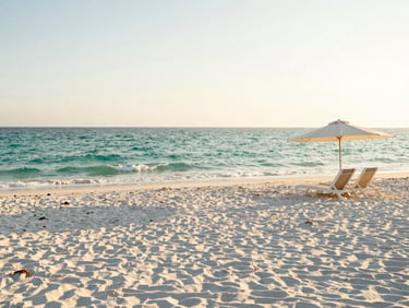 Wide panoramic view of a pristine white sand beach in Miramar Beach, gentle emerald waves, a few elegant beach chairs under an umbrella on the right, soft afternoon sun.