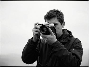 Desaturated 35mm film still of a travel vlogger holding a vintage camera, overcast daylight, high-contrast monochrome