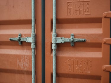 Close-up overhead shot of a shipping container door latch and seal, worn orange paint and stamped cargo markings visible under diffuse warehouse floodlight
