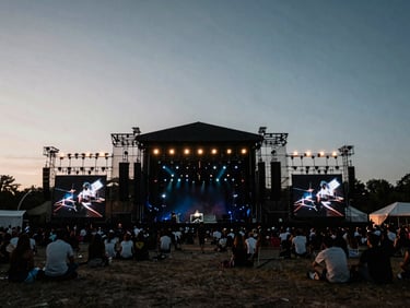 Wide environmental shot of a massive outdoor festival stage at twilight, dramatic high-contrast event lighting, deep rich shadows.