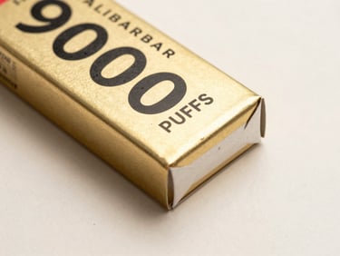 Close-up studio flat of ALIBARBAR INGOT 9000 PUFFS pack on a clean cream surface, soft diffuse overhead light, gold-foil label detail sharp, subtle rim highlight on pack edges, minimal warm-white background