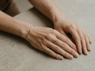 A close-up of hands gently resting on a neutral stone surface, showcasing a minimalist ring and bracelet, warm earth tones, shallow depth of field.