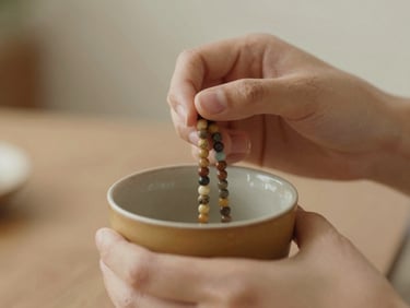 A close-up of hands holding a beaded necklace over a warm ceramic cup, soft focus background, warm earth tones.