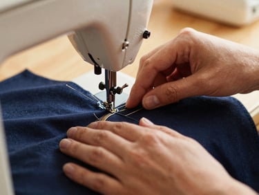 Close-up of hands working on a sewing machine, stitching structured navy fabric, warm natural light, shallow depth of field, 35mm