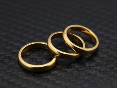 Overhead flat-lay of three identical polished gold rings on a dark carbon background, sharp highlights, 85mm lens