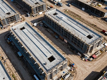 Overhead drone shot of modular construction site with completed sections and open framework sections side by side, geometric grid visible from above, clear midday light
