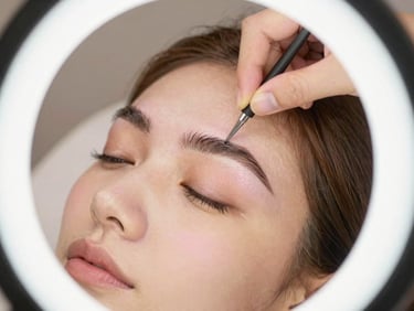 Close-up of a client receiving brow shaping with precise lines, diffused ring-light illumination, pristine surfaces, warm gold and soft pink tones.