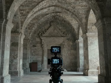 Interior of a stone monument hall, archival documentary lighting, cool and diffused, camera equipment silhouetted in foreground, vaulted ceiling visible above, atmosphere of a serious historical film production
