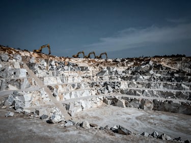 Wide shot of a massive limestone quarry, showing layers of white and grey rock, with heavy excavators in the background under an overcast sky