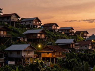 Balikpapan hillside neighborhood at dusk, timber houses on stilts visible against a warm orange sky, street lamps beginning to glow, shot from a low vantage point looking uphill