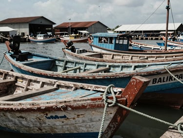 Close-up of a Balikpapan harbor at midday, wooden fishing boats moored in a row, textured rope and weathered hull in foreground, port warehouses visible behind, natural daylight, documentary framing
