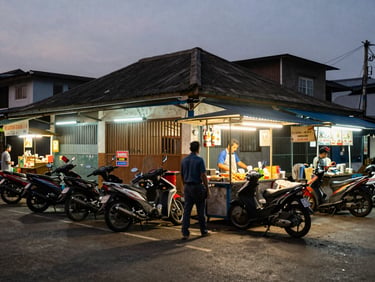 Wide shot of a Makassar street corner at dusk, food stall lit by warm fluorescent light, motorbikes parked along a shop-front row, pedestrians passing, ambient street glow, documentary style