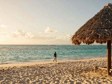 Wide environmental shot of Punta Cana coast at golden hour, a thatched palapa on the right edge, turquoise water and white sand stretching left, a traveler walking toward the water in silhouette, warm sky overhead