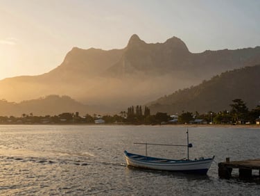 Wide environmental shot of Santa Marta bay at morning light, Sierra Nevada peaks visible in the misty background, a narrow fishing boat leaving the dock in the foreground, golden warm tones on the water surface