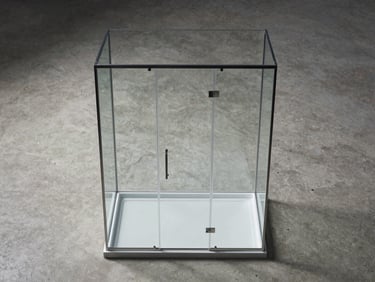 Overhead flatlay of a frameless glass shower enclosure panel and a slim pivot mampara frame on a concrete studio surface, cool diffused studio light, no people