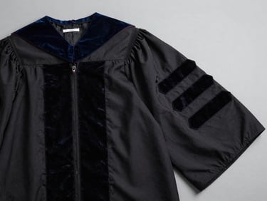 Close overhead flat-lay of a Doctorat-level academic gown with wide velvet facings and doctoral hood, black fabric on light grey surface, studio overhead light, detail on sleeve facing width