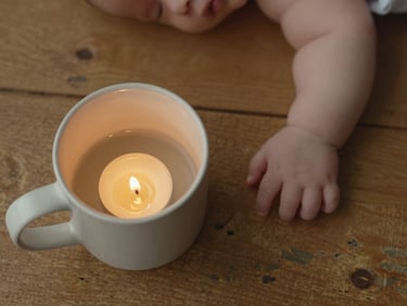 Overhead close-up of a ceramic mug on a worn wooden surface beside a sleeping infant's small hand, candlelight warmth, quiet domestic stillness, no faces