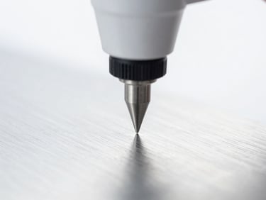 Macro shot of a surface roughness tester's stylus moving across a polished metal surface, bright clinical studio lighting, sharp focus, cool desaturated tones, 50mm