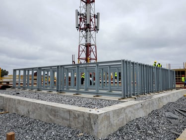 Mid-range environmental photograph of an active cell tower construction site: concrete foundation formwork in foreground, structural steel legs staged horizontally on gravel pad, overcast daylight, site crew in hard hats and high-visibility vests visible at mid-frame for scale, no portraits, cold steel and grey gravel tones