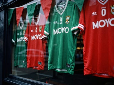 Close-up of a vintage Mayo GAA jersey hanging in a crowded Dublin pub window, green and red bunting, overcast daylight, high contrast, 35mm film.