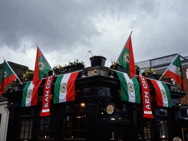 Wide shot of a London pub exterior draped in Mayo GAA flags and scarves, dramatic overcast daylight, high contrast, 35mm film.