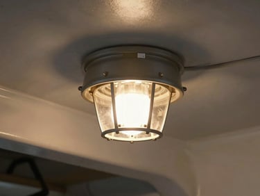 Mosquito killer lamp fixture mounted on a vessel's interior ceiling, compact industrial housing, warm tungsten ambient light from galley behind it, tight environmental framing