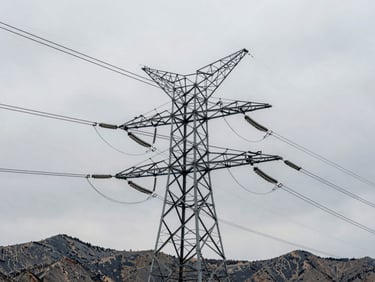 Low-angle heroic shot of high-voltage transmission towers rising against a cold, overcast Utah mountain sky, industrial steel rigging in sharp focus, desaturated tones.