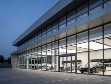 A high-contrast, dramatic architectural wide shot of a modern automotive dealership at dusk, sharp steel and clean glass reflecting cool ambient light, negative space on the left.